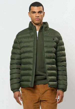Man wearing green quilted puffer jacket over green zipper sweater and brown corduroy pants, standing against plain background.