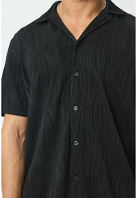 Chemise noire à manches courtes avec un motif de vagues texturé, dotée d'un col et d'une fermeture à boutons, fabriquée en tissu léger.