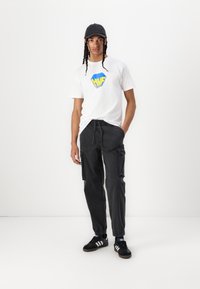Hvid t-shirt med blåt og gult logo, sorte cargo-bukser med sidelommer, sort kasket og sorte sneakers med hvide striber.