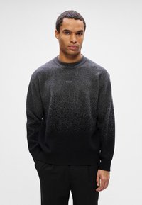 Homem vestindo um sweater em gradiente preto e cinza com o logótipo "BOSS", combinado com calças pretas, a posar contra um fundo claro e liso.