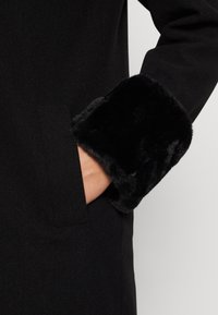 Manteau noir avec un poignet en fausse fourrure douce noire. Présente une poche latérale et un design structuré. Texture lisse, apparence élégante.