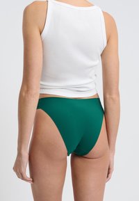 Slip bikini verde realizzato in materiale morbido e elastico. Aperture per le gambe a taglio alto e copertura minima sul retro. Design semplice e senza parti metalliche visibili.