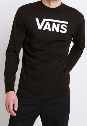 Mann trägt ein schwarzes Langarmshirt mit dem weißen Vans-Logo auf der Brust, steht mit einer Hand hinter dem Rücken.
