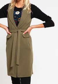 Chaleco sin mangas verde con cinturón de lazo, bolsillos de parche y cuello de solapa, llevado sobre una blusa negra de manga larga con un diseño floral colorido.