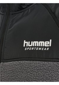 Nærbillede af sort og grå fleece sportstøjjakke med "hummel SPORTSWEAR" logo og synlige lynlåssømme.