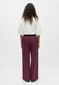 Pantalons larges rayés bordeaux avec une taille plate et deux poches arrière, associés à une chemise blanche ample. Tissu texturé.