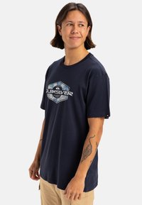 Camiseta de algodón de color azul marino con un gráfico hexagonal que presenta el texto "Quiksilver" en blanco. Diseño de manga corta y cuello redondo.