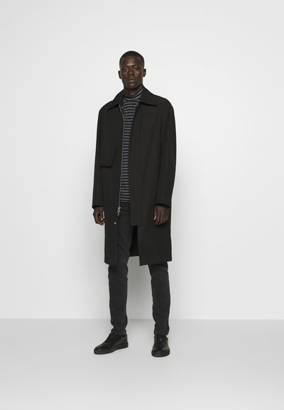 Long manteau noir avec un col replié, associé à un haut à rayures sombres, un pantalon sombre et des chaussures noires. Design minimaliste, texture lisse.