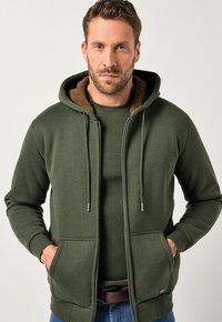 Grüne Zip-Hoodie mit einer braunen, mit Fell gefütterten Kapuze, verstellbaren Kordeln und Fronttaschen. Kombiniert mit einem dunkelgrünen Rundhals-Shirt.