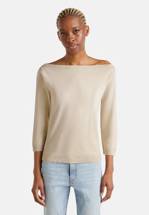 Beige gebreide top met een off-shoulder halslijn, driekwart mouwen en geribde onderrand. Draagt met lichtblauwe versleten spijkerbroek.