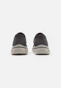 Zapatos negros tipo slip-on con parte superior texturizada, forma redondeada y suelas de color gris claro. Presentan una sutil línea blanca a lo largo del borde.