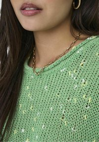 Zielony sweter dziany z fakturowanym wzorem z żółtymi i białymi kropkami. Widoczny złoty naszyjnik z wydłużonymi ogniwami.