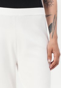 Witte joggingbroek met een geribbelde zwarte tailleband, met een zachte textuur en een slim-fit ontwerp. Hand zichtbaar met tatoeages.