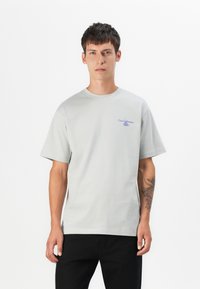 Hellgraues Baumwoll-T-Shirt mit kurzen Ärmeln, Rundhalsausschnitt und einem kleinen lila Grafikdruck mit der Aufschrift "Casa Garbana" auf der linken Brust.
