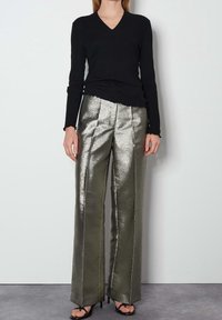 Svart långärmad V-ringad topp i kombination med högmidjade, metalliska olivgröna byxor som har vertikala sömmar och en flytande, glänsande textur.