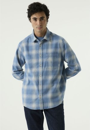 Mann mit kurzen, lockigen Haaren trägt ein blau-weiß kariertes Hemd und dunkle Jeans, stehend vor einem schlichten, hellen Hintergrund.