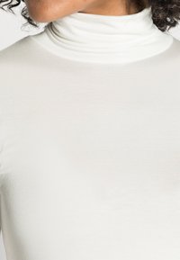 Nahaufnahme einer Person, die einen weißen gerippten Rollkragenpullover trägt, mit sichtbaren lockigen Haaren und Hautfarbe am Hals und Kiefer.