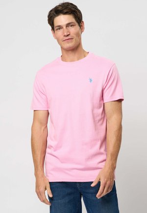 Moški stoji, oblečen v svetlo rožnato majico s kratkimi rokavi z modrim logotipom na prsih in temno modre kavbojke, na enobarvni svetli podlagi.