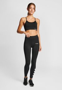 Zwarte atletische beha met dunne bandjes, gecombineerd met hoge zwarte leggings met een zijpatroon en logo. Draagt zwarte sneakers.