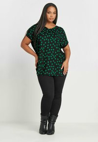 Mustat paita, jossa on vihreitä pilkkuja, lyhyet hihat ja V-pääntie. Yhdistetty tiukkoihin mustiin farkkuihin ja polveen asti ulottuviin mustiin saappaisiin.