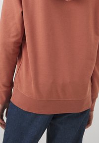 Persona con un sudadera color óxido y pantalones azul oscuro, vista desde atrás, con una mano parcialmente visible.