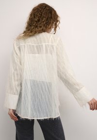 Translucent white blouse with a textured, hairy finish. Long sleeves and a loose fit. Features a hip-length design with a straight hem.