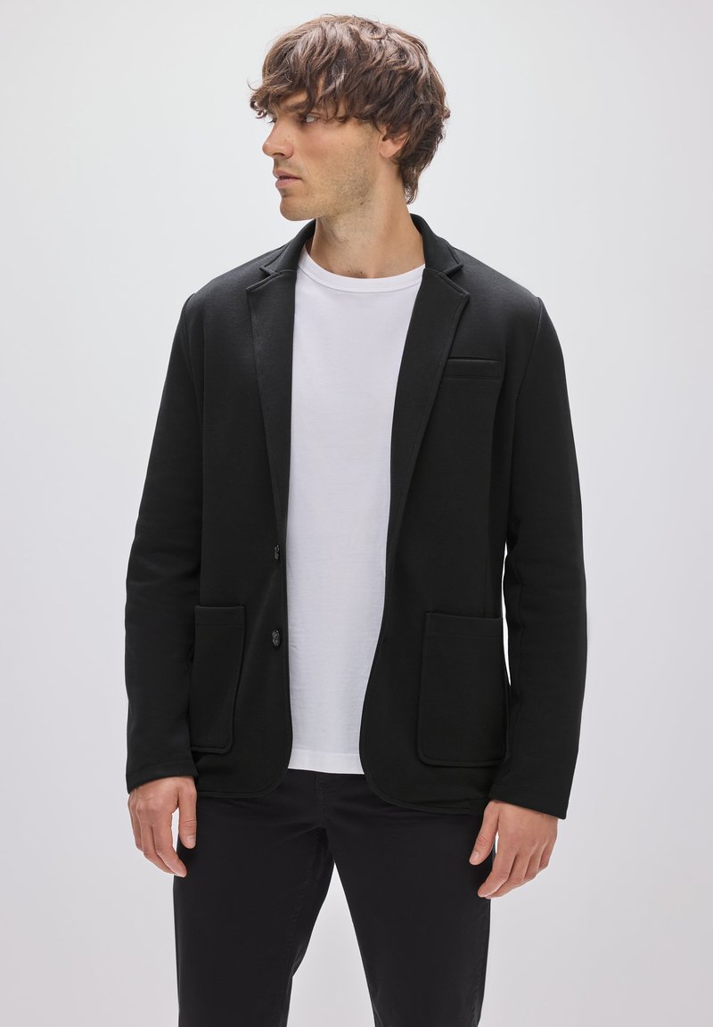 Blazer negro de tejido estructurado, con un botón único, dos bolsillos frontales y un bolsillo en el pecho; llevado sobre una camiseta blanca y pantalones negros.