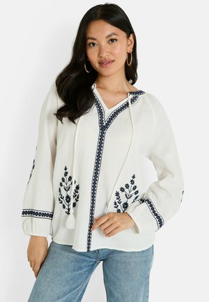 Blouse blanche à manches longues avec broderie florale bleu marine et décolleté en V, dotée de cordons de serrage et de poignets froncés. Tissu doux et texturé.