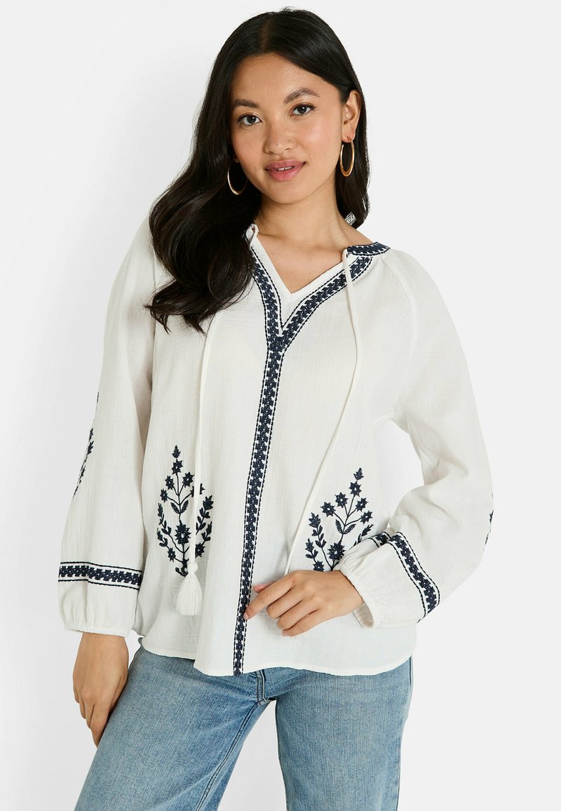 Blouse blanche à manches longues avec broderie florale bleu marine et décolleté en V, dotée de cordons de serrage et de poignets froncés. Tissu doux et texturé.