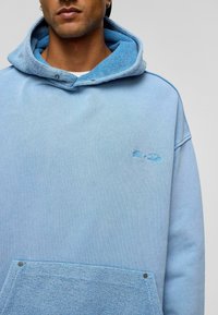  Lichtblauwe hoodie met een gestructureerde capuchon, voorzien van een ruime zak en een subtiel logo. Het materiaal lijkt zacht met een ontspannen pasvorm.