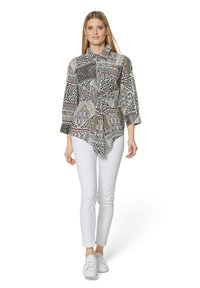 Blaue Bluse mit einem Kragen, die eine Mischung aus schwarzen, weißen und bunten geometrischen Designs aufweist. Kombiniert mit weißen Hosen und Sneakers.