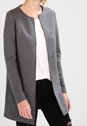 Femme portant un long cardigan gris texturé, ouvert sur l’avant, par-dessus une chemise blanche et un jean noir déchiré, debout devant un fond uni.