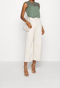 Zielona bluzka z koronkowymi detalami, w zestawie z białymi spodniami o szerokich nogawkach. Akcesoria obejmują małą srebrną torebkę na ramię i cieliste szpilki.