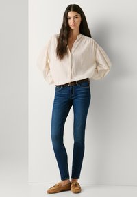 Γυναίκα με μακριά σκούρα μαλλιά, που φοράει εκρού μπλούζα, σκουρό μπλε στενά τζιν, καφέ ζώνη και καφέ μοκασίνια, στέκεται μπροστά σε έναν λευκό τοίχο.