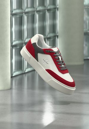 Λευκό, κόκκινο και γκρι sneaker Calvin Klein με λογότυπο στο πλάι, αιωρούμενο πάνω από ένα αντανακλαστικό γκρι πάτωμα με φόντο τοίχο από γυάλινα τουβλάκια.