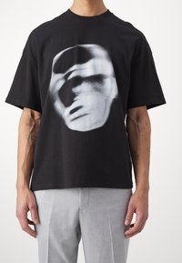 Black cotton t-shirt featuring an abstract white face graphic, short sleeves, and a relaxed fit. Paired with light gray pants.