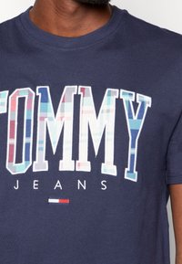 Camiseta de algodón azul marino con la palabra "TOMMY" en letras grandes y coloridas de cuadros y "JEANS" en blanco, con un pequeño emblema rojo, blanco y azul.