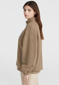 Beige színű fleece pulóver magas nyakkal, cipzáros záródással és kontrasztos barna zsebbel. Jellemzője a lelógó váll és az elasztikus mandzsetta.