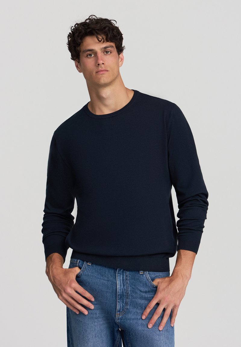 Maglione girocollo blu navy in morbido tessuto a maglia con polsini e orlo a coste, abbinato a un paio di jeans di media tonalità, in piedi su uno sfondo neutro.