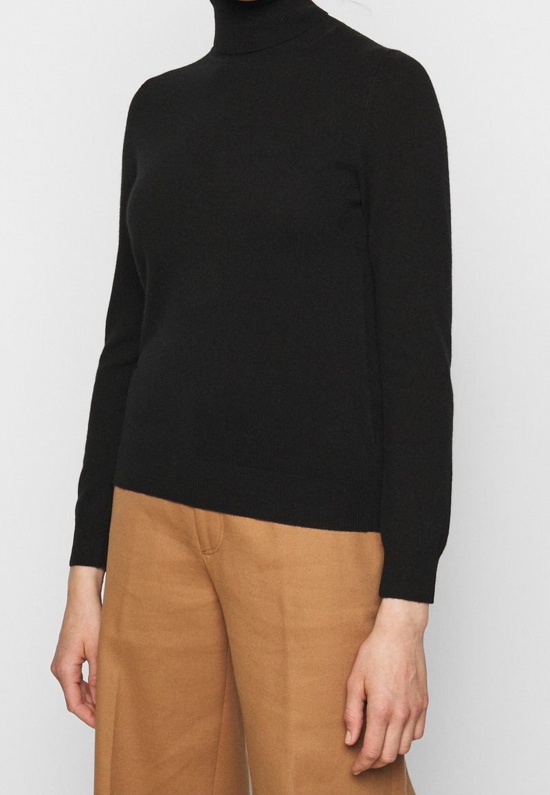 Pull noir à col roulé à manches longues associé à un pantalon large taille haute de couleur camel sur une personne debout.