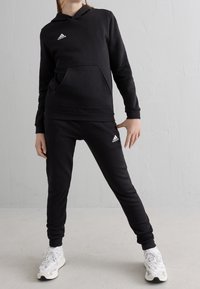 Conjunto de sudadera con capucha y pantalones deportivos negros hecho de tela suave. Cuenta con un bolsillo frontal y detalles de logotipos blancos en el pecho y la pierna del pantalón.