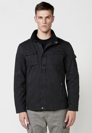 Black fabric jacket featuring a high collar, multiple front pockets, and a button closure, with a textured finish and side details.