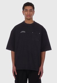 Sort oversized t-shirt lavet af bomuld, med rund hals, korte ærmer og en brystlomme med knapdetalje. Hvid tekstdesign.