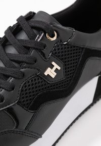 Baskets de sport noires avec une tige en mesh texturé, accents en cuir élégant, œillet doré et une semelle blanche présentant un motif de design prononcé.