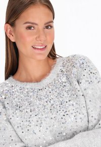 Szürke, bolyhos pulóver, kerek nyakkivágással, színes flitterekkel díszítve, amelyek texturált és csillogó felületet alkotnak a mellkason.