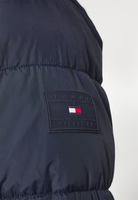 Marinblå puff jakke med matte overflate, som har en rektangulær svart lapp merket "TOMMY HILFIGER" og små røde, hvite og blå detaljer.