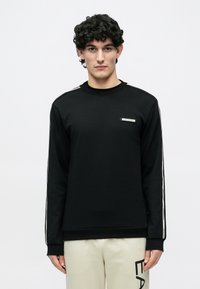 Jauns vīrietis ar cirtainiem matiem, valkā melnu garām piedurknēm džemperi ar baltām sānu svītrām un gaiši bēšas bikses ar melniem burtiem.