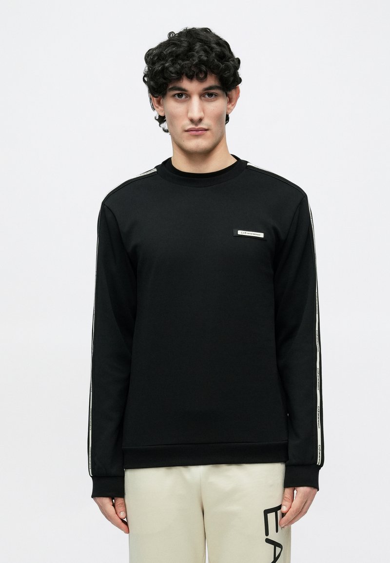 Jauns vīrietis ar cirtainiem matiem, valkā melnu garām piedurknēm džemperi ar baltām sānu svītrām un gaiši bēšas bikses ar melniem burtiem.