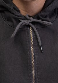 Zwarte hoodie met een textuurafwerking, voorzien van een gouden rits en grijze trekkoord. Het ontwerp heeft een lichte kraag en ribstof.