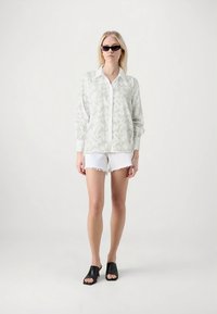 Jachetă cămașă gri, ușoară și texturată, cu guler alb, asortată cu pantaloni scurți din denim alb cu margini destrămate și mule negre cu vârf deschis.